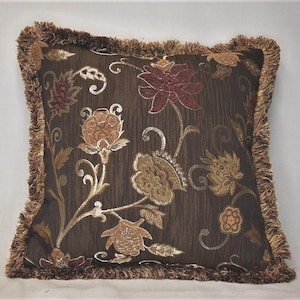 Puede incluir: Una almohada de tiro marrón con un patrón floral en tonos dorados, rojos y naranjas. La almohada tiene un borde con flecos.