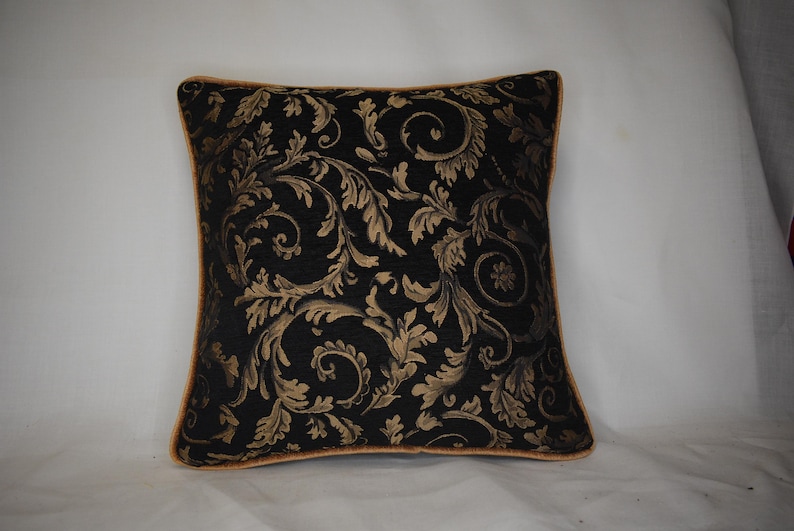 May include: Square cushion with a black backdrop and a gold floral design. The cushion has gold edging. The pattern features swirling leaves and floral motifs. Suitable for home decor, this cushion measures approximately 40cm x 40cm.