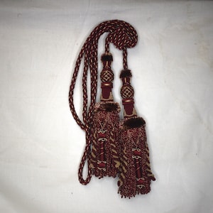 May include: A pair of burgundy tassel tiebacks with gold accents. The tassels are made of a soft, woven fabric and have a fringe detail. The tiebacks are connected by a burgundy and gold rope.