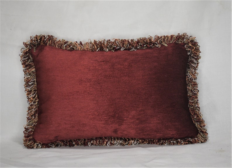 May include: A burgundy pillow with a fringed border. The fringe is a mix of brown, beige, and white.