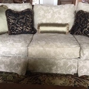 May include: A cream sofa with a floral pattern, adorned with decorative cushions. The cushions feature gold, black, and olive green hues, with diverse textures and designs. The sofa rests on a patterned rug.
