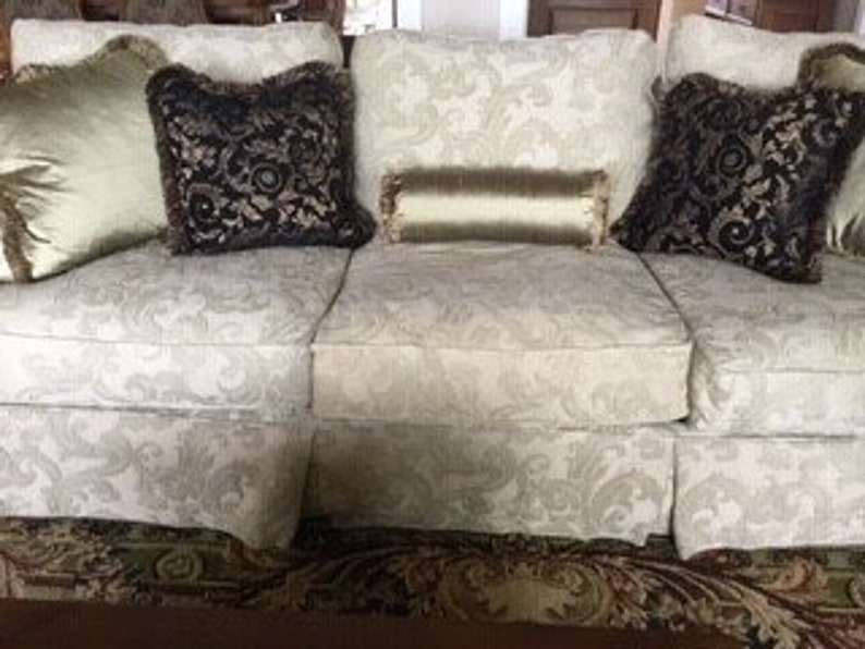 May include: A cream sofa with a floral pattern, adorned with decorative cushions. The cushions feature gold, black, and olive green hues, with diverse textures and designs. The sofa rests on a patterned rug.