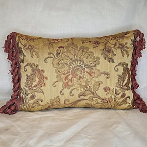 May include: Rectangular decorative pillow with a floral pattern in shades of gold, brown, and red. The pillow has a fringe trim in a deep red color. The design features a large central floral motif and smaller floral and paisley elements.