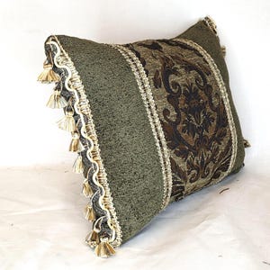 May include: A square decorative pillow featuring a textured olive green fabric panel and a central vertical band of dark brown damask pattern. The edges are trimmed with a cream-colored wavy gimp braid and a series of small hanging tassels.