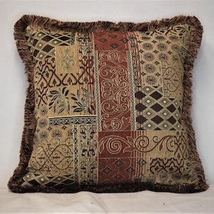 May include: A decorative throw pillow with a brown, beige, and red patterned fabric. The pillow has a fringed border and features a patchwork design with various floral and geometric motifs.
