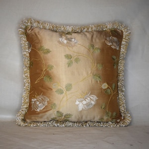 Puede incluir: Una almohada decorativa con un patrón floral dorado y crema. La almohada tiene un borde con flecos.
