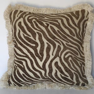 May include: A square throw pillow featuring a brown and cream zebra stripe pattern. The pillow is finished with a thick, cream-colored fringe trim along all four edges, providing a textured border to the patterned fabric surface.