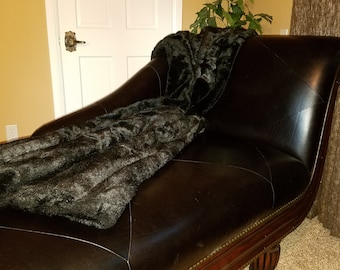 May include: A black faux fur throw draped over a brown leather chaise lounge with a wooden frame.