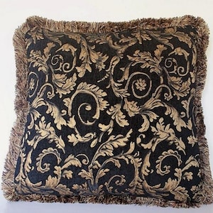 May include: Square decorative pillow with a black background and a gold floral pattern. The pillow has a brown fringe trim around the edges. The design features swirling leaves and ornate details, suitable for home decor.