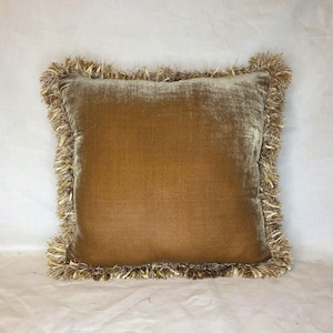 May include: A square throw pillow featuring a smooth, golden-brown velvet fabric on the front surface. The edges of the pillow are finished with a thick, textured fringe trim in a matching metallic gold and brown color palette.