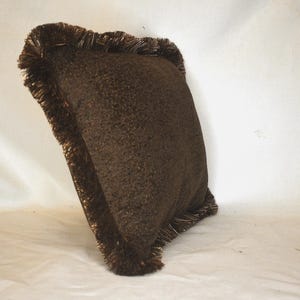 May include: A brown throw pillow with a fringe border. The pillow is made of a soft, textured fabric.