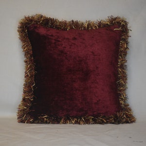 May include: A square burgundy velvet pillow with a decorative fringe border. The fringe is a mix of gold and brown tones, creating a textured edge around the pillow. The pillow is a solid color and appears soft.