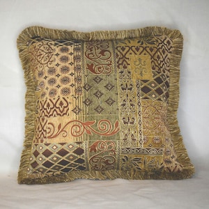 May include: A square throw pillow featuring a patchwork design of various woven fabrics in shades of gold, olive green, and dark brown. The surface displays intricate geometric, floral, and scroll patterns, finished with a thick gold fringe trim.