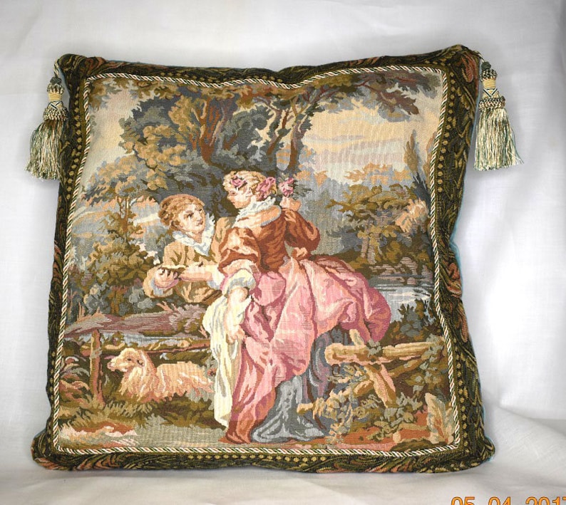May include: A tapestry pillow with a green, brown, and blue border. The pillow features a scene of two people in a forest setting. The woman is wearing a pink dress and the man is wearing a brown jacket. There is a small dog in the foreground.