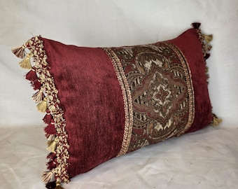 Cojín lumbar de chenilla color burdeos o óxido con flecos de borlas doradas para sofá o cama de salón.