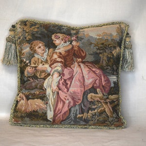 May include: A tapestry pillow with a green, brown, and pink design featuring two people in a forest setting. The pillow has a green fringe border and tassels on the corners.