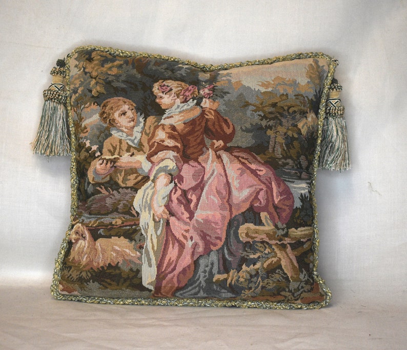 May include: A tapestry pillow with a green, brown, and pink design featuring two people in a forest setting. The pillow has a green fringe border and tassels on the corners.