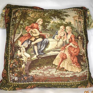 May include: A tapestry pillow featuring a scene of three people in a garden setting. The people are dressed in 18th-century clothing and are sitting on a stone bench. The man is playing a stringed instrument, while the two women are listening. The background is a lush green garden with trees and flowers.
