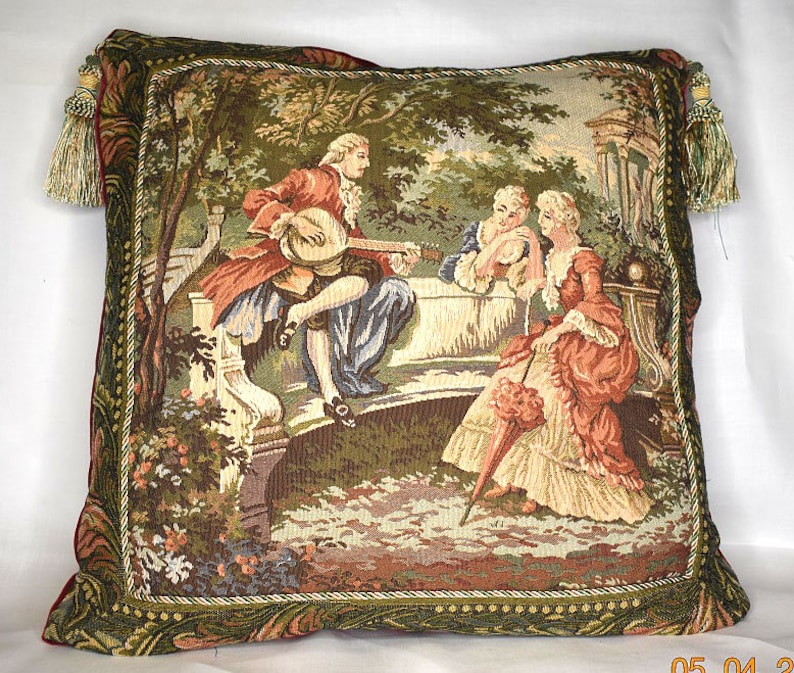 May include: A tapestry pillow featuring a scene of three people in a garden setting. The people are dressed in 18th-century clothing and are sitting on a stone bench. The man is playing a stringed instrument, while the two women are listening. The background is a lush green garden with trees and flowers.