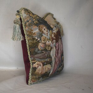 May include: A tapestry pillow with a burgundy velvet backing and gold trim. The front of the pillow features a scene of two people in a forest setting with a dog.