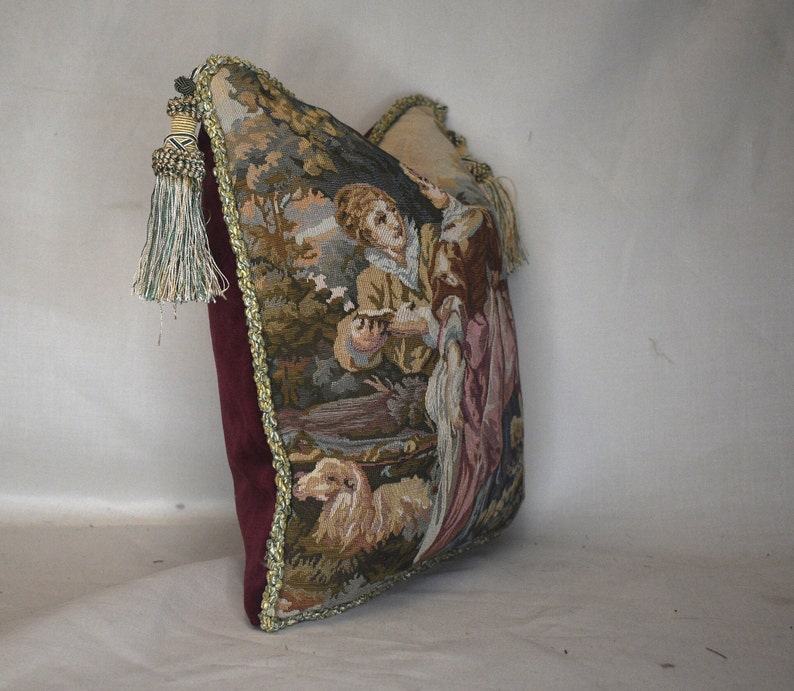 May include: A tapestry pillow with a burgundy velvet backing and gold trim. The front of the pillow features a scene of two people in a forest setting with a dog.