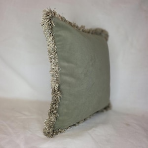 May include: A square sage green decorative pillow with a soft, velvety texture. The edges are trimmed with a thick, textured fringe in a neutral beige color. The pillow is designed for home decor.
