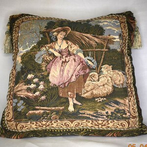 May include: A tapestry pillow with a green, brown, and beige background featuring a woman in a pink and white dress standing in a field with sheep. The pillow has a gold and brown border with tassels.