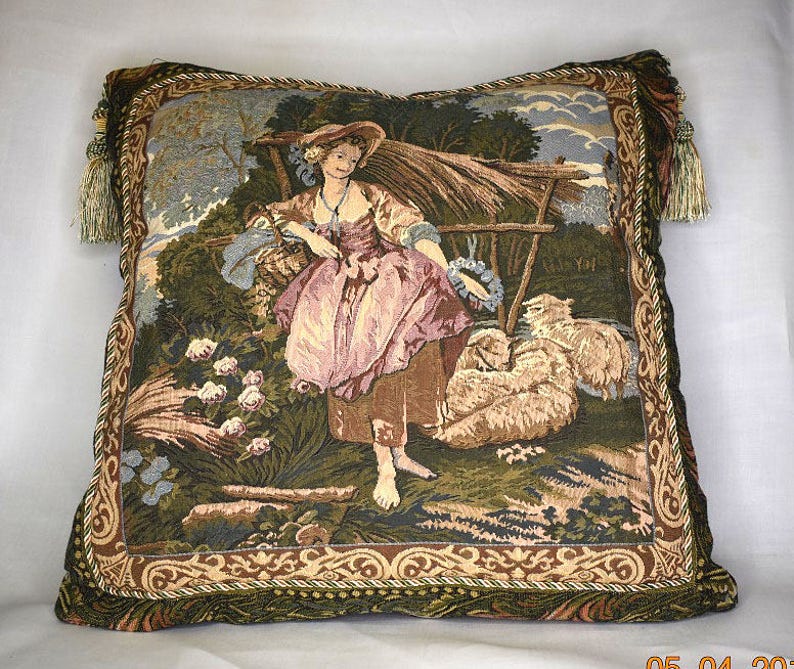 May include: A tapestry pillow with a green, brown, and beige background featuring a woman in a pink and white dress standing in a field with sheep. The pillow has a gold and brown border with tassels.