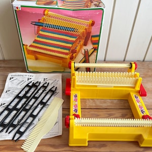 May include: A yellow plastic toy loom with two white rotating bars, accompanied by four black plastic shuttles, a beige plastic comb, and an instruction sheet. The set is presented with its original cardboard box featuring a colorful graphic.