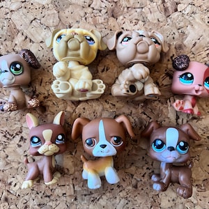 May include: A collection of seven toy dog figurines in various poses. The dogs are primarily brown, tan, and pink, with large, expressive eyes and glitter accents. The figurines are on a textured, cork-like surface.