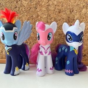 Puede incluir: Tres figuras de poni de plástico colocadas una al lado de la otra. El poni de la izquierda es azul claro con cuerpo azul oscuro, alas y una cresta roja y amarilla. El poni central es blanco con detalles y crines rosas. El de la derecha es azul oscuro con una máscara blanca.