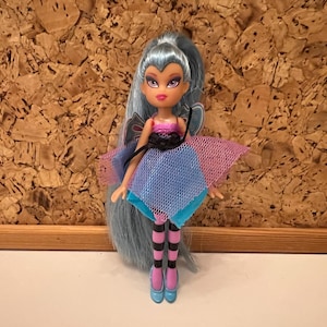 May include: A doll with long, light blue hair and a pink and black top. The doll has a blue and pink skirt, striped black and pink stockings, and blue shoes. The doll has a fairy wing design on the back.