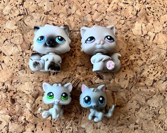 May include: Four small, plastic cat figurines in various poses. The cats are primarily grey with different eye colours, including blue and green. Some have black markings. The figurines are on a textured, brown surface.