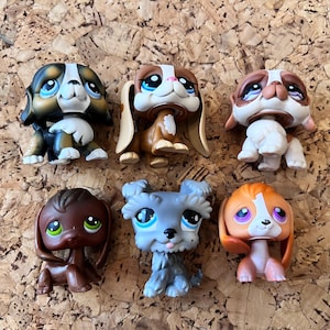 May include: Six colorful, collectible dog figurines with large eyes. The dogs are in various poses and colors, including brown, black, orange, and gray. The toys are made of plastic and are on a textured surface.