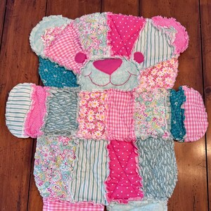 Pode incluir: Um urso de peluche de patchwork feito com tecido rosa, verde e branco. O urso tem uma borda franjada e um rosto sorridente.