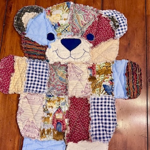 May include: A patchwork quilt teddy bear with a blue and white checkered pattern, red and white floral patterns, and a beige background. The bear has a blue nose and black eyes.