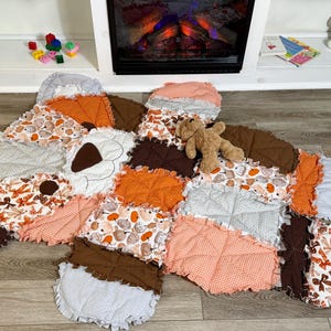 May include: A handmade rag quilt featuring a patchwork design with various autumn-themed fabrics in shades of orange, brown, and white. The quilt displays patterns of pumpkins, leaves, and gingham, with frayed edges and a small brown teddy bear resting on top.