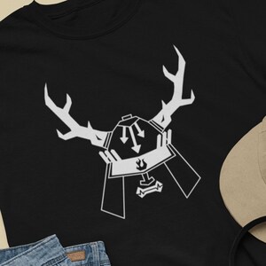 May include: Black t-shirt with a white graphic of a samurai helmet with antlers. The graphic has two arrows pointing down. The t-shirt is laid flat on a light brown surface with a pair of blue denim shorts, black sneakers, a brown baseball cap, and a skateboard.