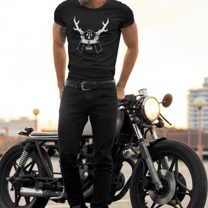 May include: A man wearing a black t-shirt with a white graphic of a samurai helmet with antlers. He is standing next to a black motorcycle.