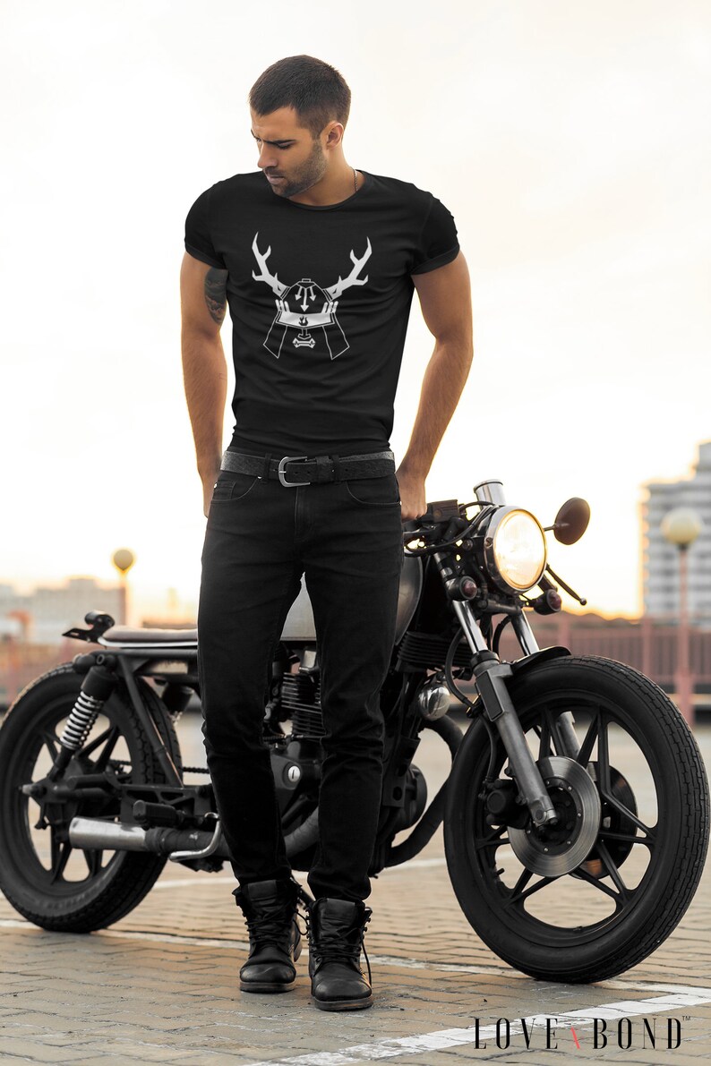 May include: A man wearing a black t-shirt with a white graphic of a samurai helmet with antlers. He is standing next to a black motorcycle.