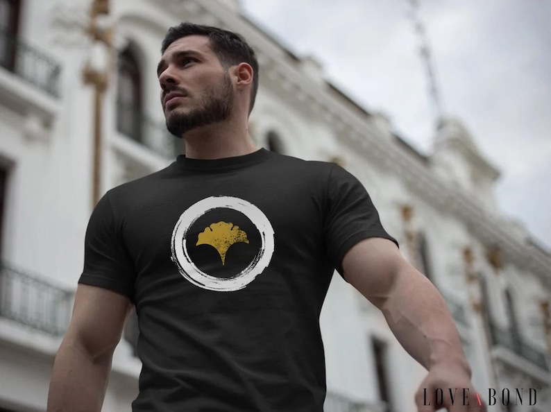 Pu&ograve; includere: Una maglietta nera con un cerchio bianco e un design a foglia di ginkgo dorata. Il testo "LOVE & BOND" &egrave; visibile nell'angolo in basso a destra dell'immagine.
