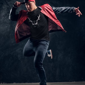 May include: A man wearing a black t-shirt with a white deer graphic, a red and black bomber jacket, blue jeans, and a red baseball cap. He is posing with his arm extended and his other hand on his cap.