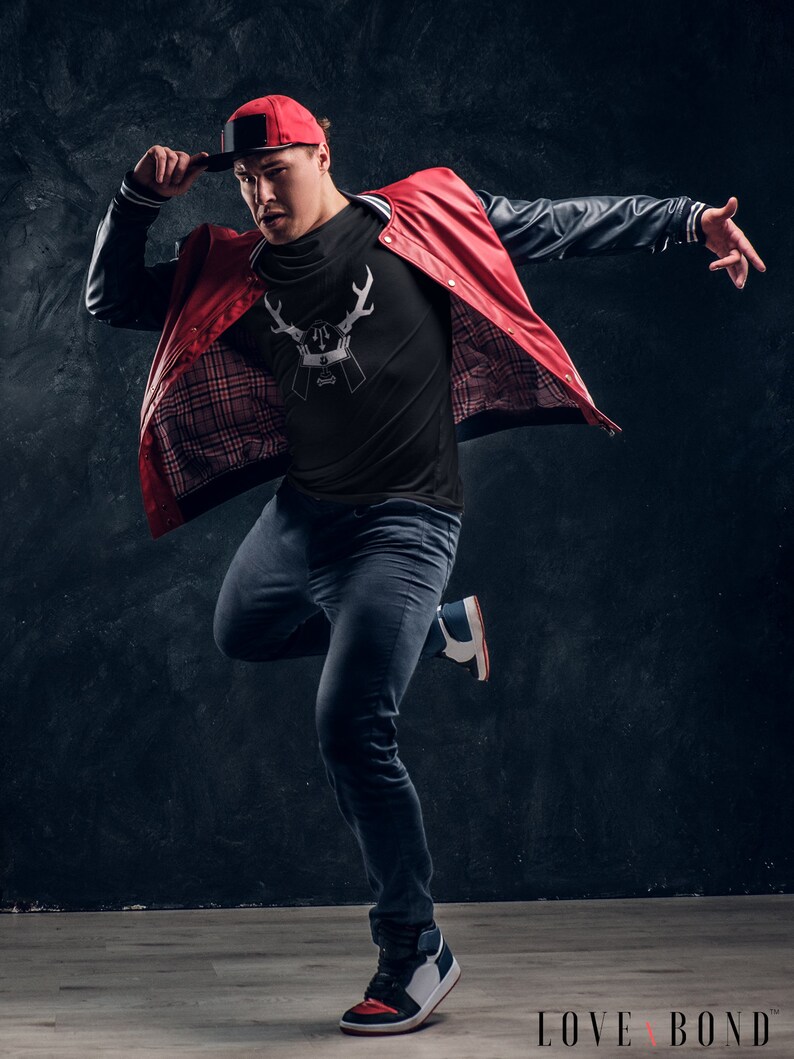 May include: A man wearing a black t-shirt with a white deer graphic, a red and black bomber jacket, blue jeans, and a red baseball cap. He is posing with his arm extended and his other hand on his cap.