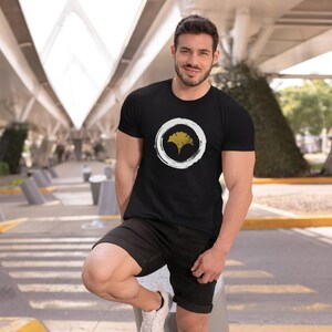 Pu&ograve; includere: Una maglietta nera con un cerchio bianco e un design a foglia di ginkgo dorata. Il cerchio ha un effetto a pennellate.