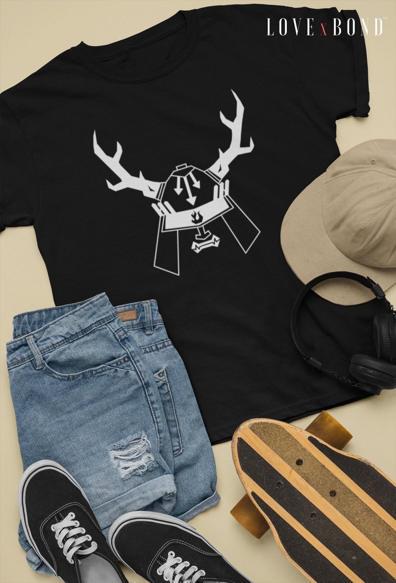 May include: Black t-shirt with a white graphic of a samurai helmet with antlers. The graphic has two arrows pointing down. The t-shirt is laid flat on a light brown surface with a pair of blue denim shorts, black sneakers, a brown baseball cap, and a skateboard.