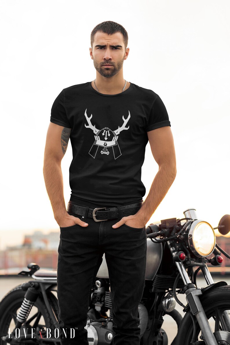 May include: A black t-shirt with a white graphic of a samurai helmet with antlers. The man is wearing black jeans and a black belt. He is standing in front of a motorcycle.