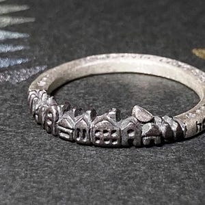 May include: A silver ring featuring a miniature cityscape design. The ring has a band with a series of tiny houses and buildings, creating a skyline effect. The ring is on a dark surface.