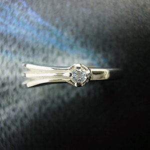 May include: A silver ring with a single, clear gemstone set in the center. The ring has a unique, curved design.
