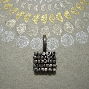 May include: A silver charm with a square design made of small, round beads. The beads are arranged in a pattern of crescent moons.