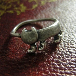 May include: A silver ring shaped like a cat with four legs and a tail. The ring is sitting on a red leather surface.